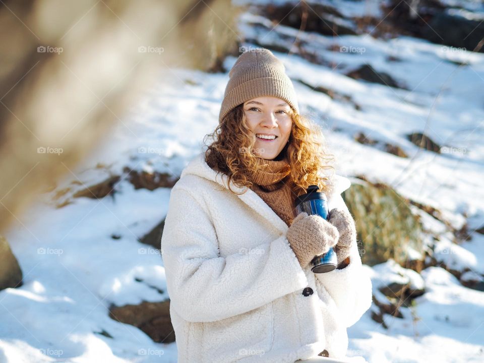 Young happy woman with curly hair in fur coat and hat drinking coffee at forest in winter 