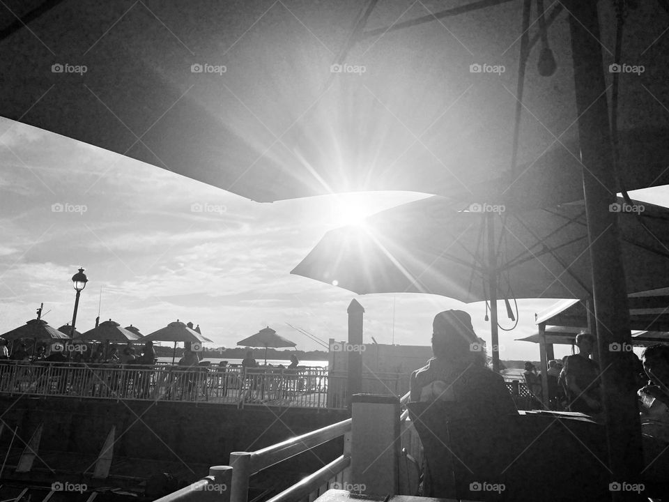 A black and white image of people dining at a bar/restaurant on the 9th Avenue Pier in Belmar, NJ. It’s a very relaxed place to eat and have a cocktail while looking at the water and the boats. The sun shines down through the umbrellas.