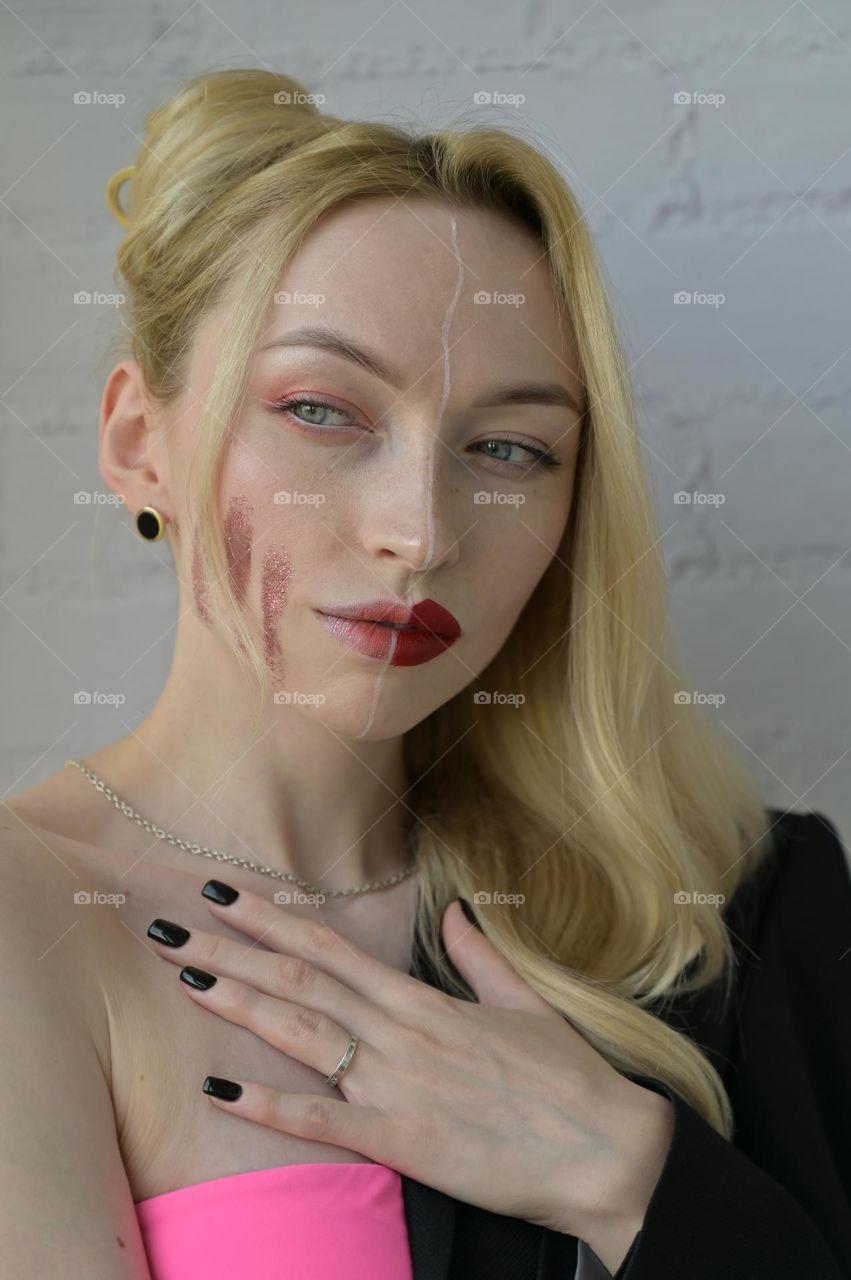 Close-up portrait of a blonde girl with different make-up on half of her face and a thoughtful look with hand on the chest on white background