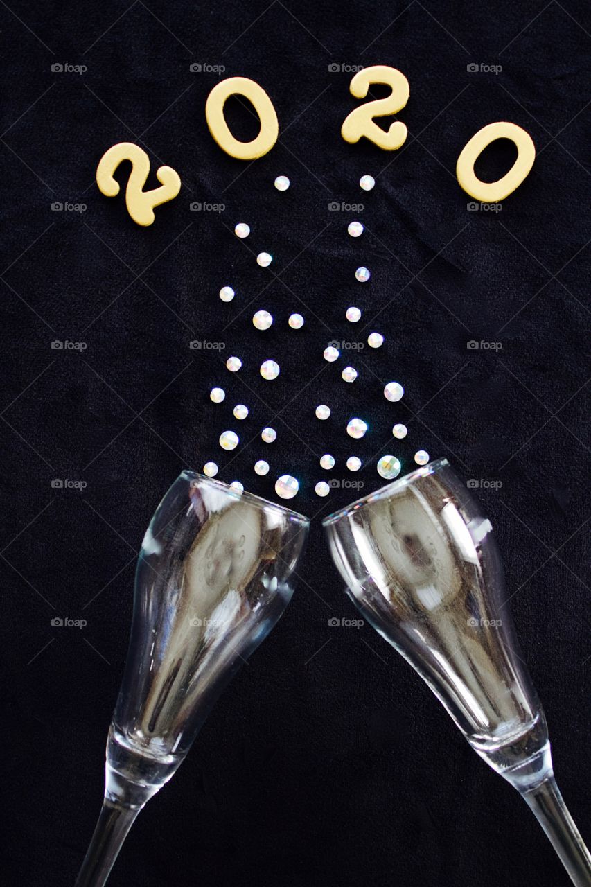 Flat lay of the numbers ‘2020’ in gold above gleaming champagne glasses with rhinestone bubbles on black background - portrait format