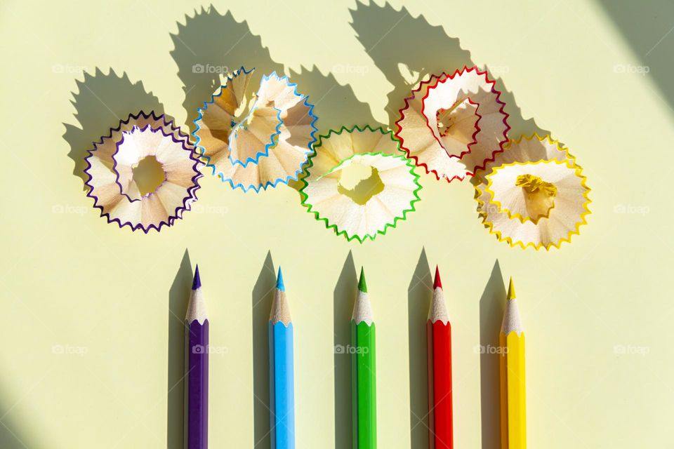 Five bright, colored pencils on a yellow background with wooden shavings and a drop shadow.