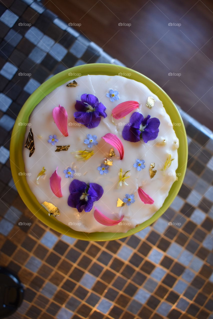 A small round cake is decorated with pink, purple and light blue flowers and gold leaf, sitting on a green cake stand. 