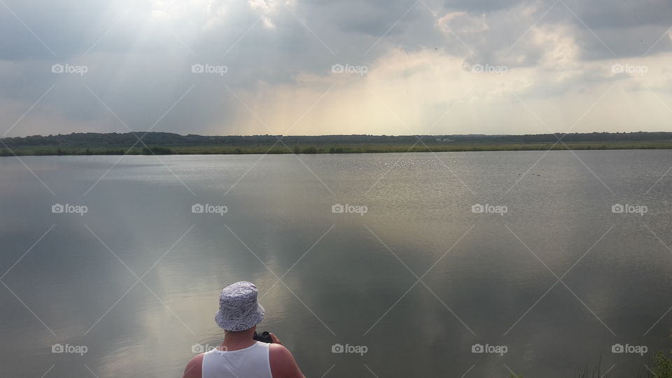 Man Wearing  a hat looking over a large lake With beautiful sky - water landscape - holding binoculars - clouds - Overcast