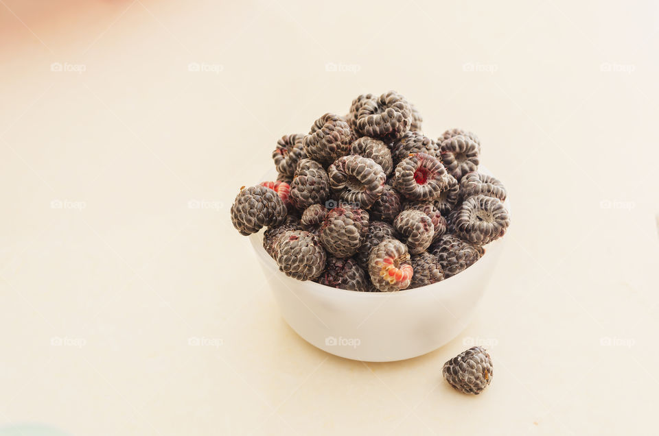 Ripe black Raspberry in white dish on a white tile countertop. One Raspberry beside the dish.