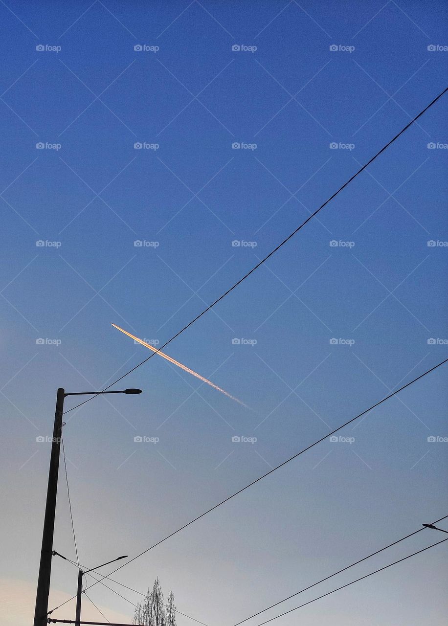 A beautiful photo of the clear blue sky, a plane flying and leaving a white streak behind and street lamps with wires in Bulgaria