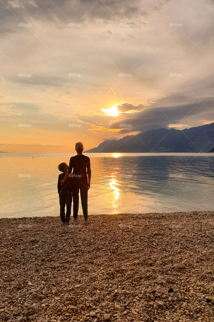 Mom with a baby boy stand embracing on the seashore and watching the sunset.  Summer family vacation at sea