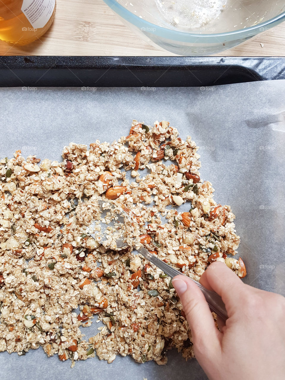 Hand making homemade granola in an oven tray on a kitchen countertop with an empty salad bowl and pot of honey
