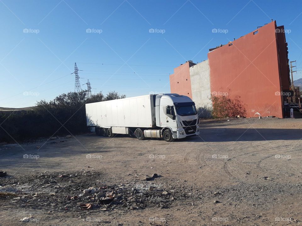 A white semi-truck is parked on a dusty lot near a large red building in Marrakech. The background features power lines, bushes, and a clear blue sky. The scene reflects an industrial or transport setting. January 1, 2025