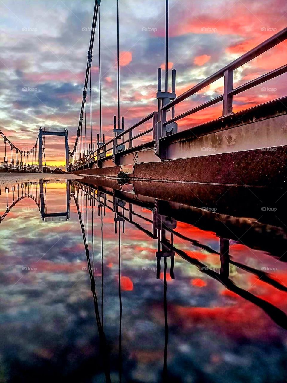 Side view of Lorois bridge in Plouhinec and its large puddle into which reflect the sunrise's grey, yellow and red colours explosion and the bridge's metal structure with at the far end, the bridge bathed in a gradient of grey, yellow and orange