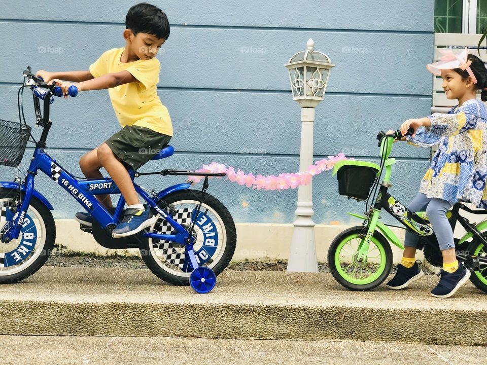 A boy on cycle helping a girl cycle to pull forward with a bunch of flower garland.😜