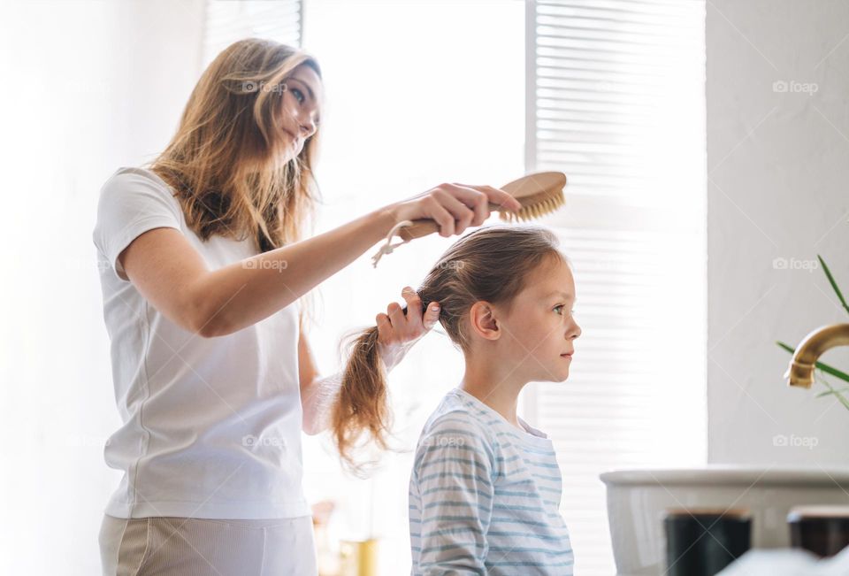 Young mother woman with long hair with little tween girl daughter in pajamas combs her hair on morning in bathroom at home