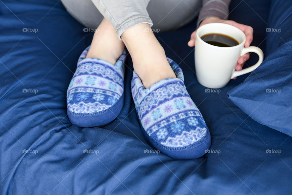 Close-up of a woman's slippered feet crossed on a bed and holding a cup of coffee