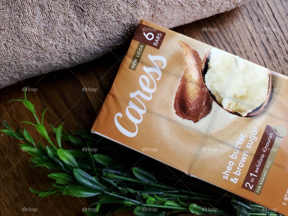 The new look Caress bar soap shea butter and brown sugar six pack,  next to a tan towel and accented with greenery on the left,  on a wood surface.