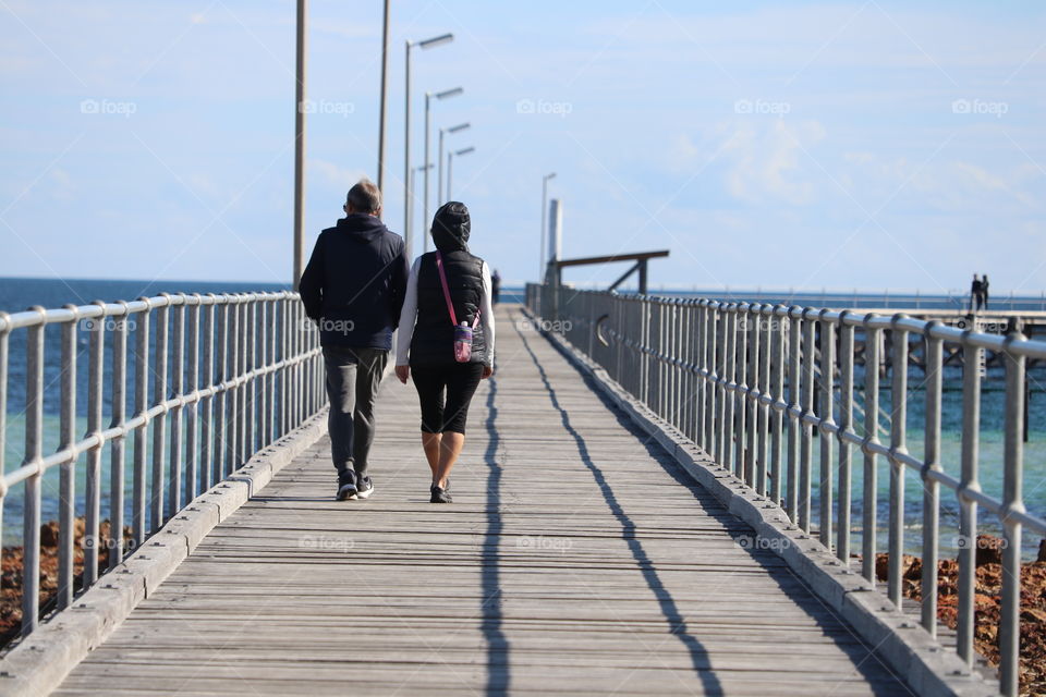 Couple male female walking down public dock wharf Jetty on sunny day ocean side 