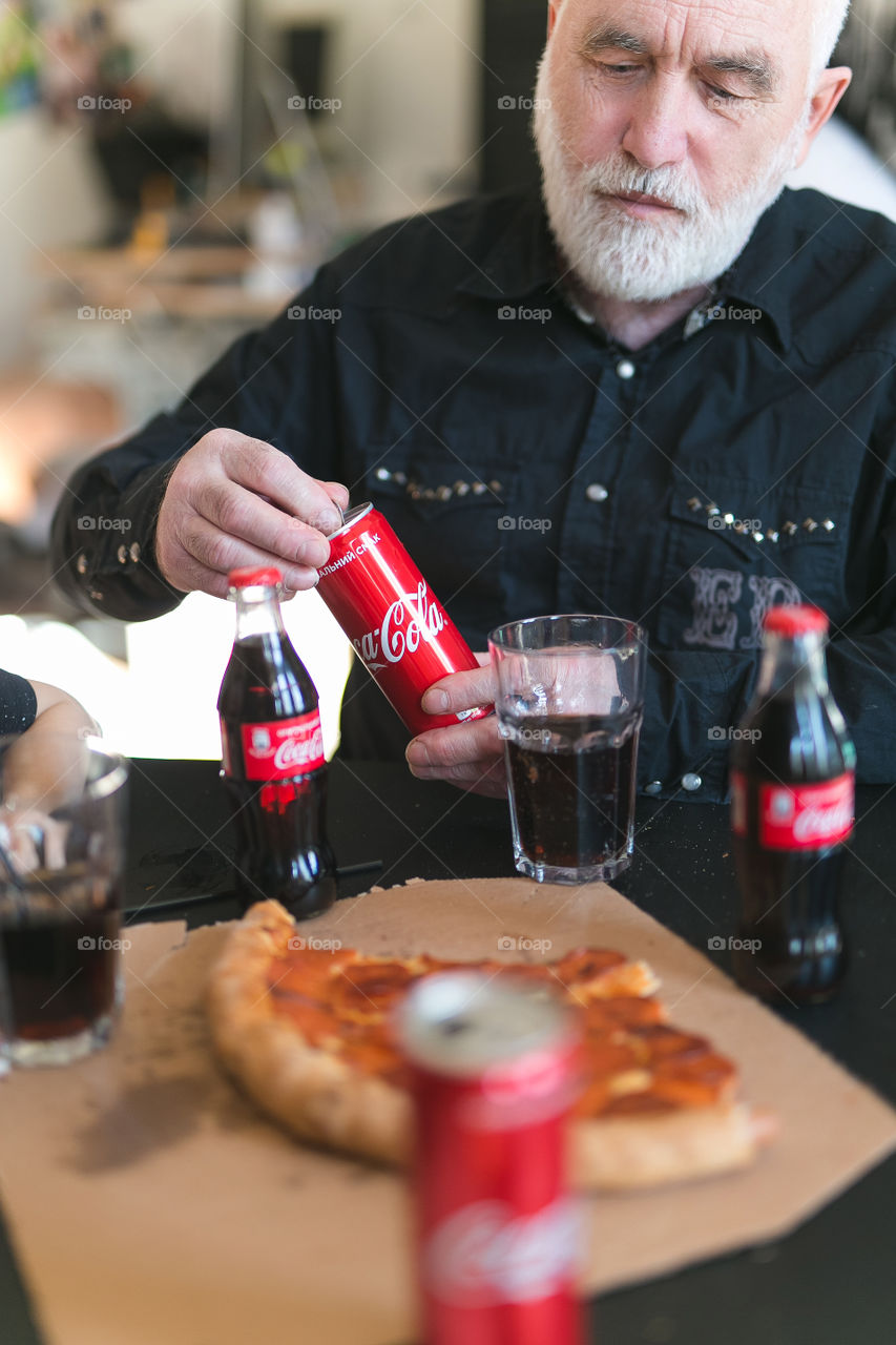 a sixty-year-old man eats lunch at home, drinks Coca-Cola, the man wears a sulfur shirt, has a gray beard and hair