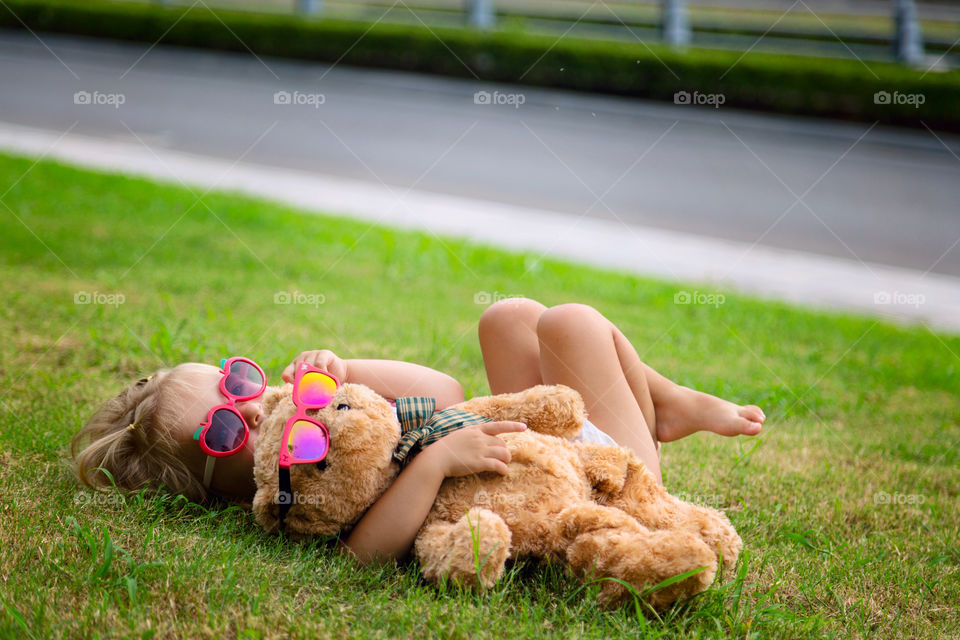 Cute little girl with blonde hair in sunglasses lying on the grass and embracing her teddy bear 