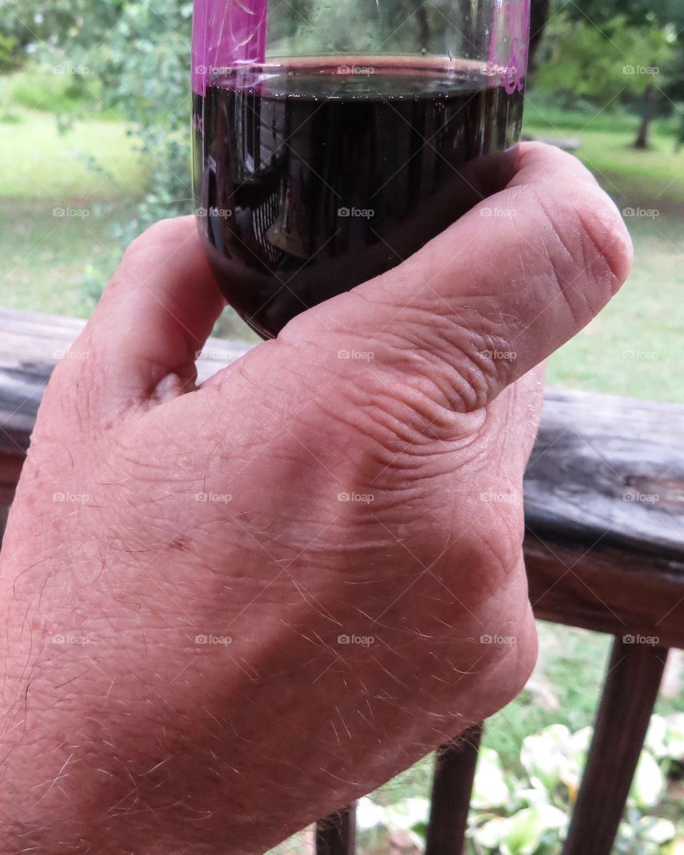 Older man holding a glass of red wine while sitting on the porch.

