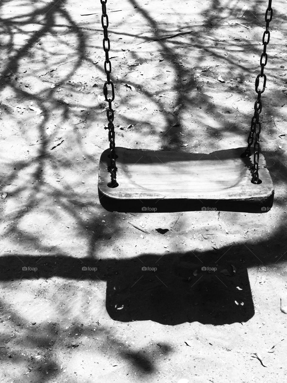 A wooden swing in a small sandy playground near the beach in Puerto Escondido, Mexico is shaded by trees with fallen leaves. The bright afternoon sun is creating shadows of the tree branches in the sand surrounding the swing.