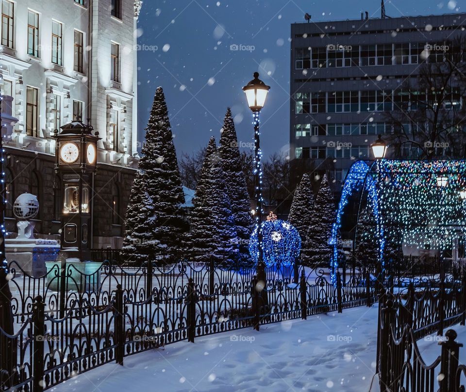 City winter night landscape. Square with a clock tower and snow-covered Christmas trees