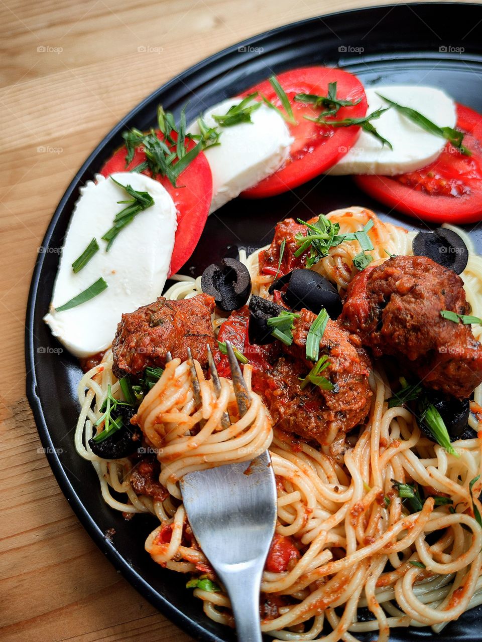 A plate of pasta, meatballs, slices of mozzarella and a sliced ​​tomato.  In the foreground, a fork with pasta