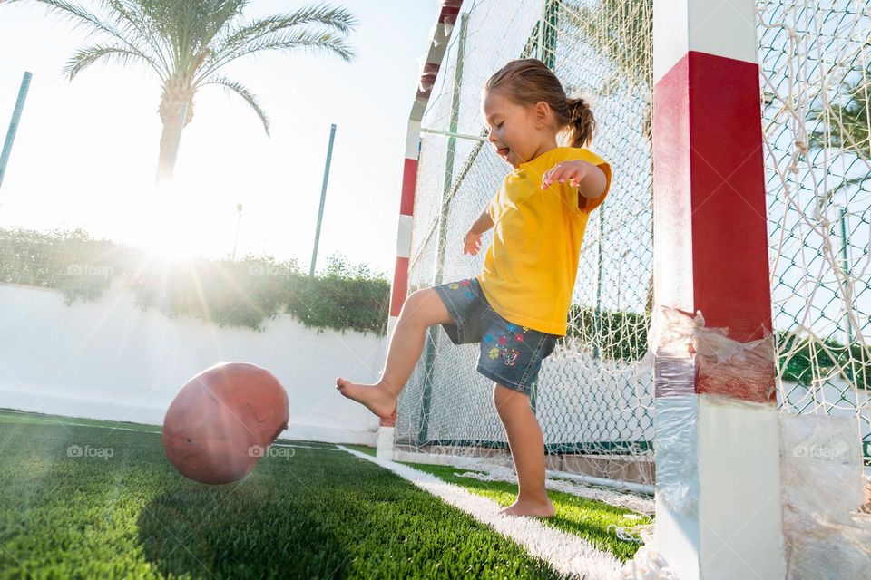 Cute little girl kicking soccer ball on green sports field at sunset time. Kid playing football with her family