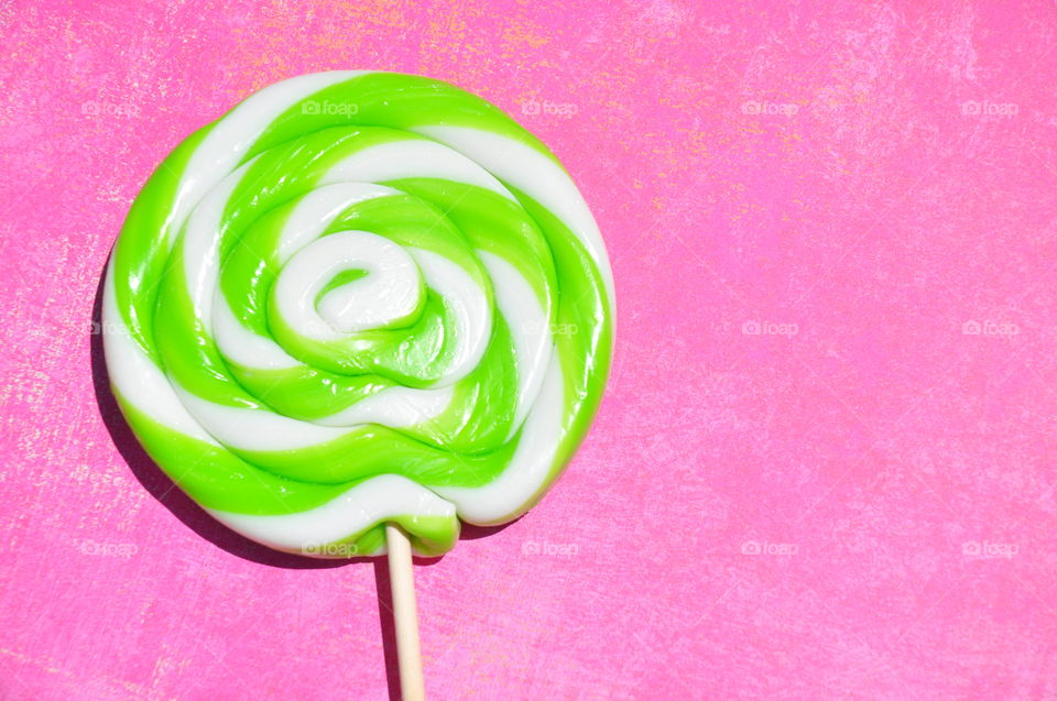 Foap mission Sugar: A large round green and white swirled lollipop against a bright pink background. 