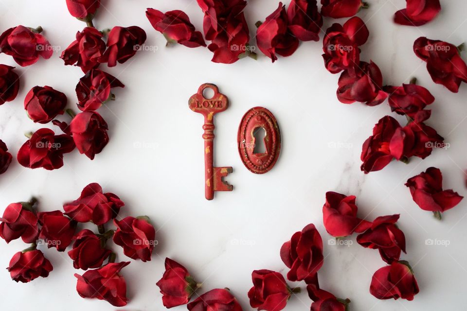 Red, metal vintage key and keyhole with red, silk rose buds in a heart-shaped arrangement on white marble surface