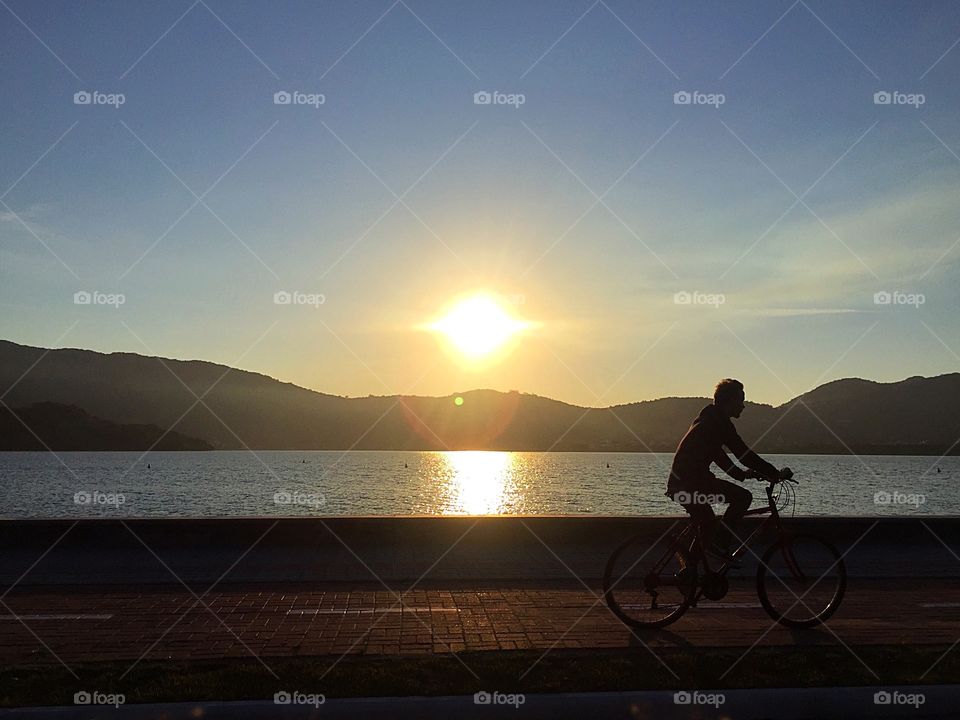 Pôr-do-sol na orla da Lagoa da Conceição em Florianópolis (SC), enquanto um ciclista aproveita a linda paisagem para um passeio ao fim da tarde. Que lugar maravilhoso! 