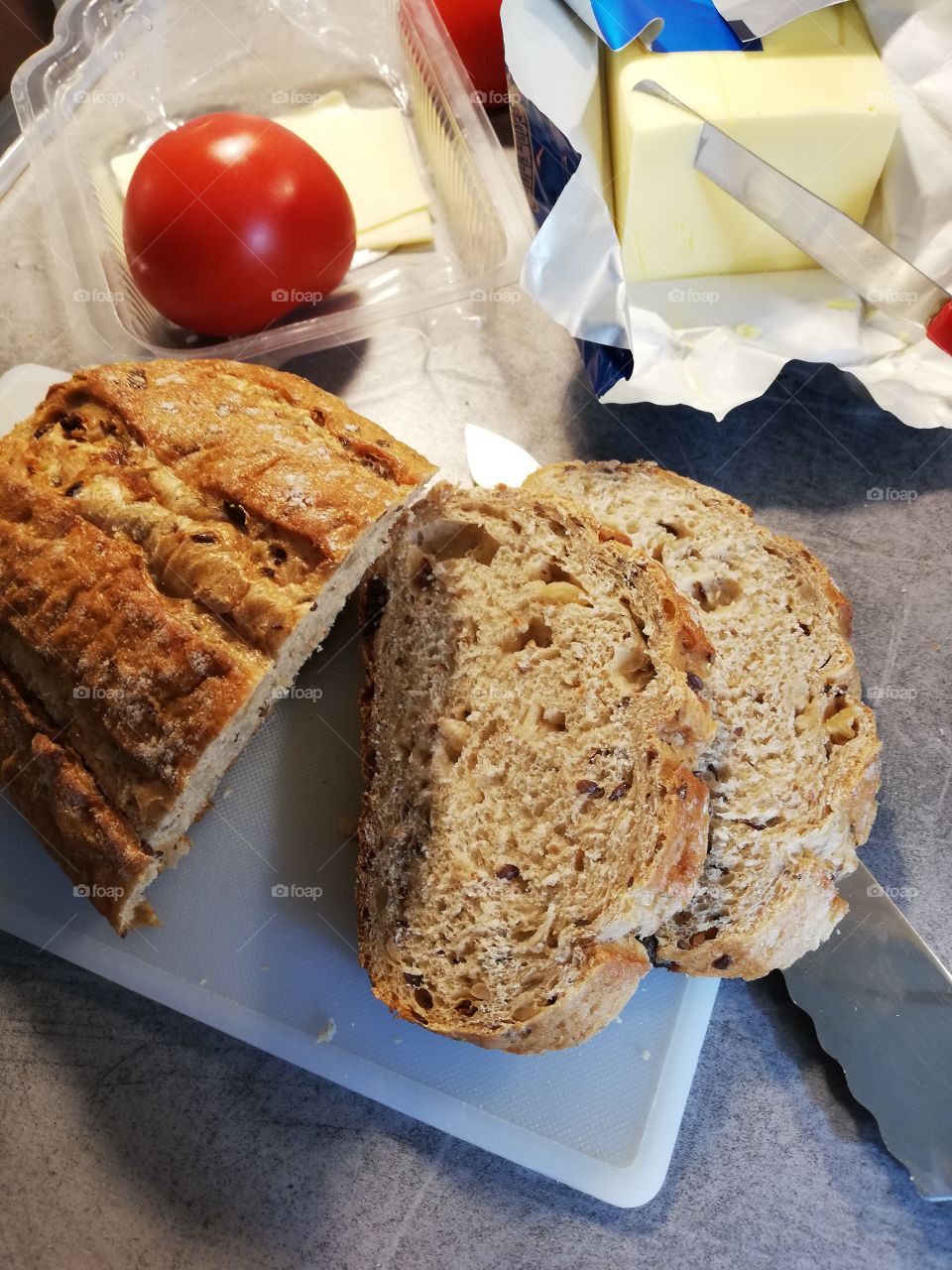 A loaf of bread with fruit and seeds on a chopping board, some crumbs on it. An edge of a knife is wavy. In an open butter package is another knife with red handle. A tomato and cheese slices in a plastic box.