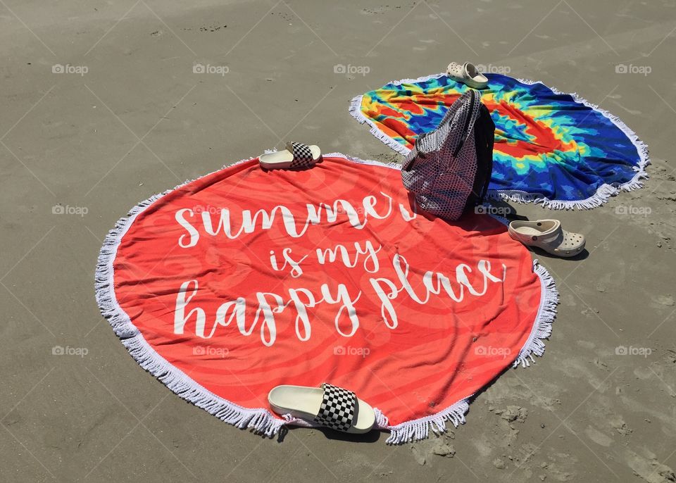 Colorful round beach blankets, one tie dyed, the other bright orange with “summer is my happy place”, on the beach secured in place with shoes and a backpack 