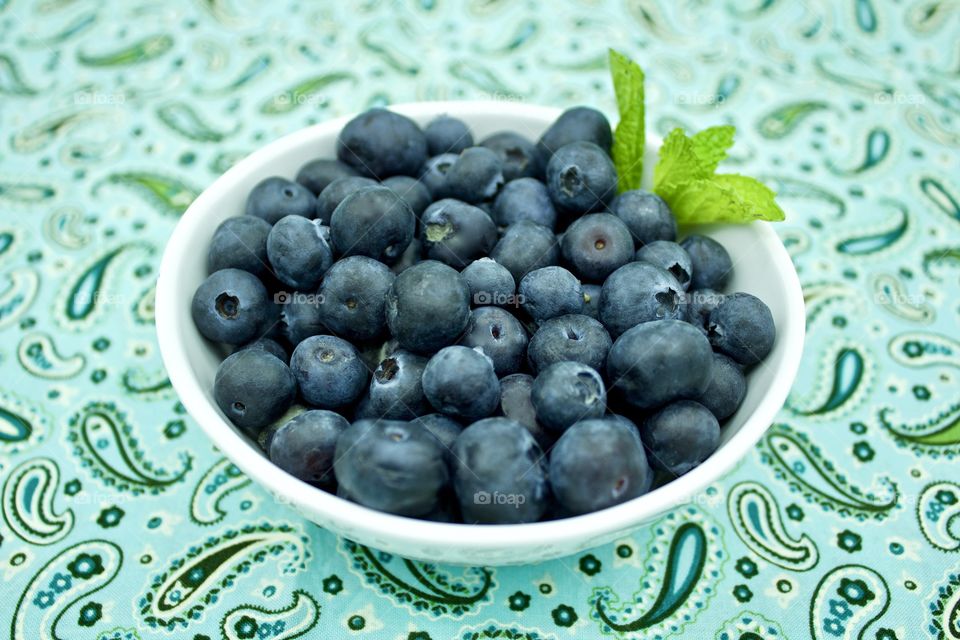 Blueberries and a spring of mint in a white bowl on teal and green paisley fabric