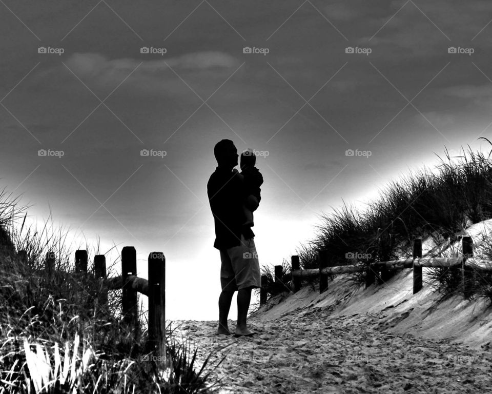 A father holds his baby boy on a walkway to the beach as the sun rises 