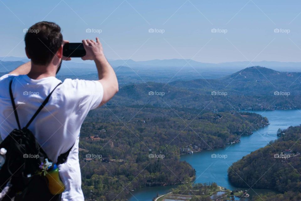 image of man on top of a mountain taking a cell phone photo of the breathtakingly beautiful scenery of the valley and lake below