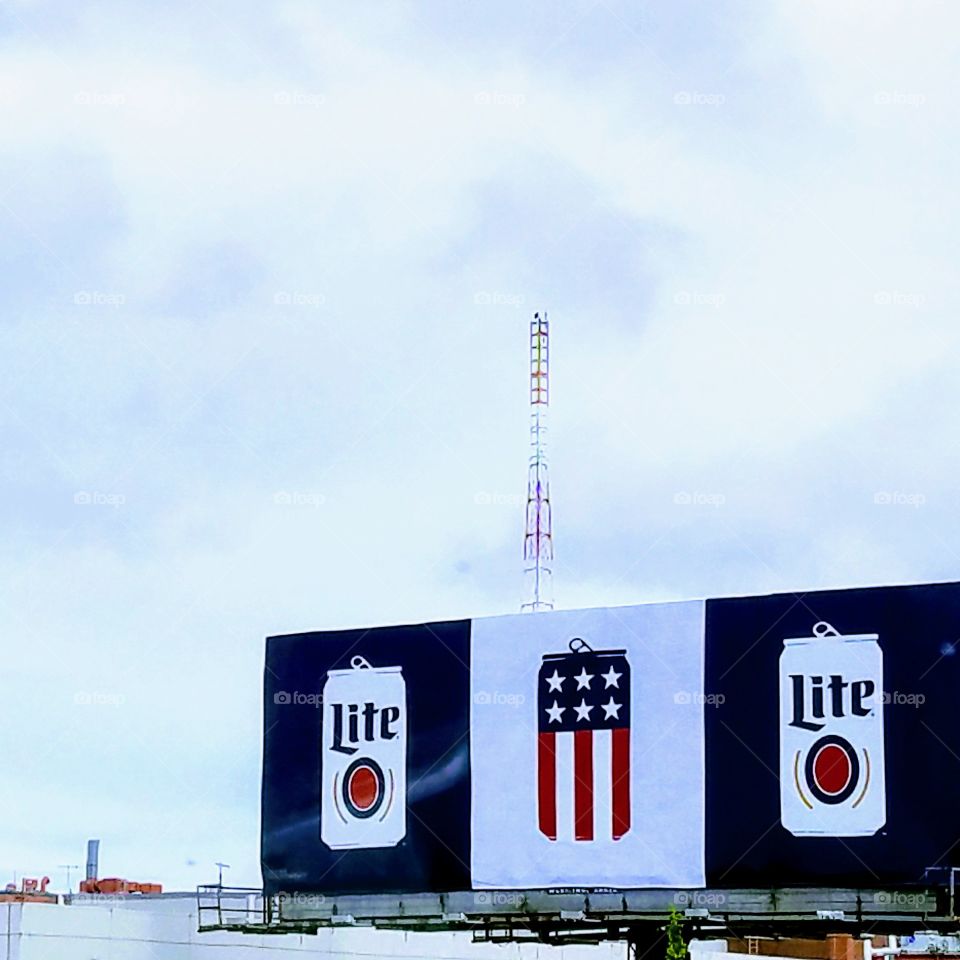 Lite Beer cans, USA flag colors on a large billboard sign. Tall tower in background on bright day.