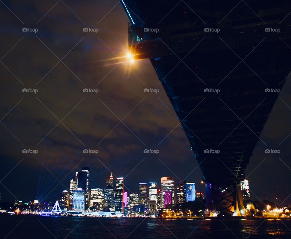 Looking up at the Sydney harbour bridge as the lights on the tall buildings surround the water 