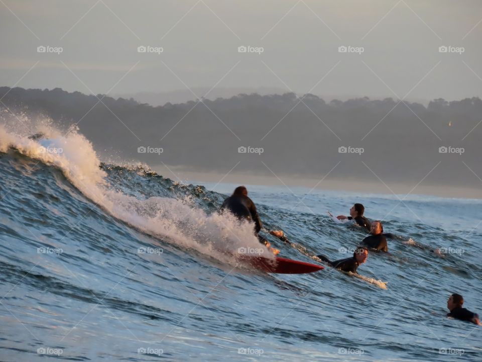 Summer by the ocean: surfer gliding down a wave at sunset as other surfers watch on.