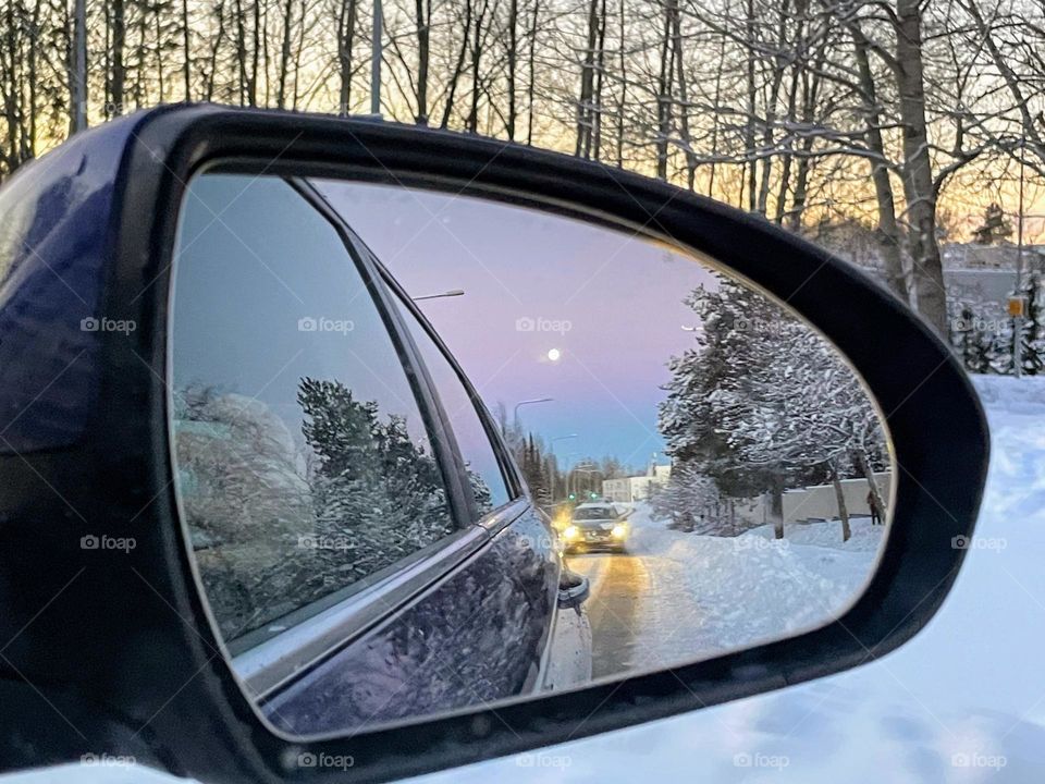  View in the car side mirror, snowy landscape, road with driving behind cars, trees and full moon on the blue purple sky 