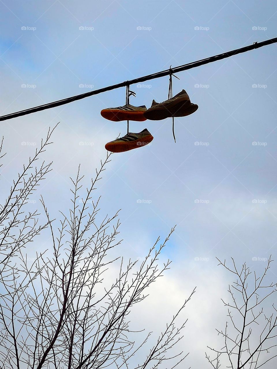 Two pairs of sneakers hang on an electric wire against the background of bare trees and a cloudy sky