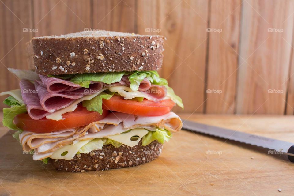 mixed meat sandwich with lettuce and tomatoes piled high on grain bread served next to a bread knife on a wood table with a wood background