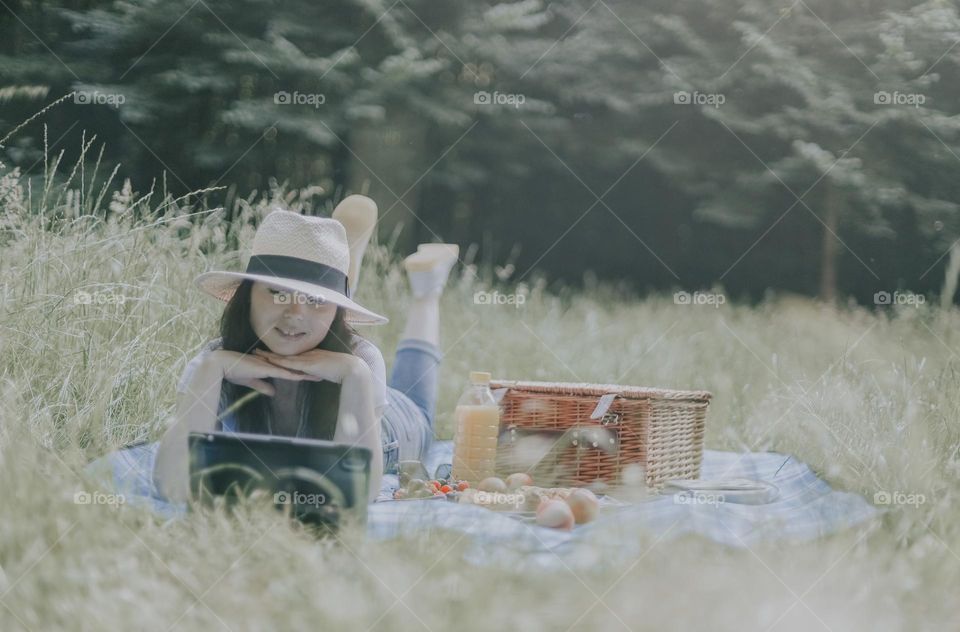 One Young beautiful caucasian girl in a straw hat lies on a bedspread with a basket of books, food, drinks and watches a movie on a tablet with her chin tucked in her hands in a meadow with tall grass on a summer day, close-up view from below.