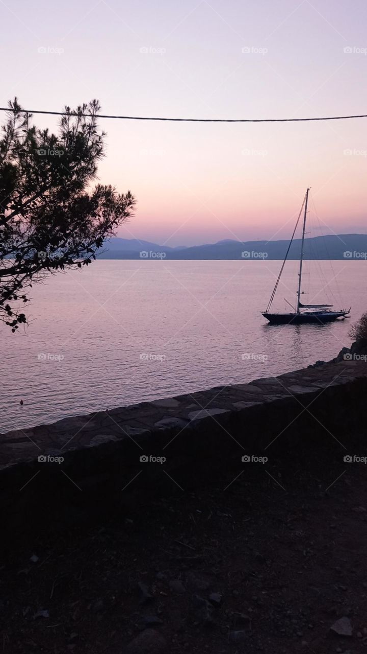 A beatyful and colorful sunset in Hydra island, Greece. In the middle of the calm sea there is a sailing boat dropping anchor for the night.