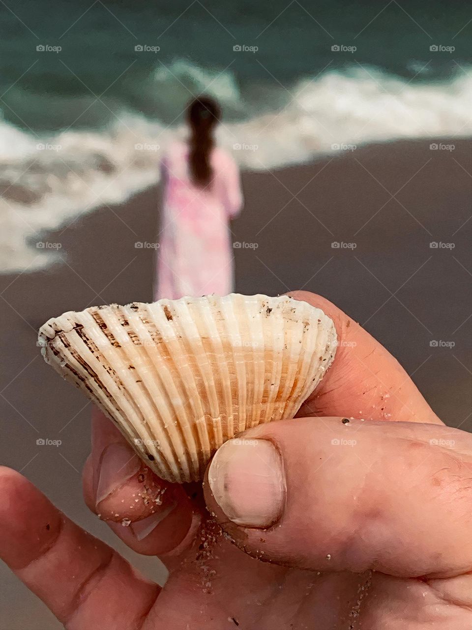 Girl dressed in pink on vacation at the beach seems to be standing inside a seashell held by a hand with a toe instead of a thumb on the hand at the shore by the waves.