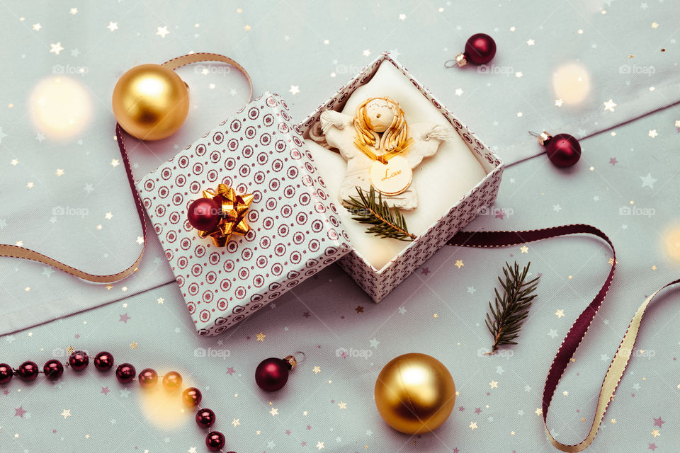 Wrapping Christmas gift into box. Decorating using red ribbon, red Christmas balls and pine twigs. Blurred Christmas lights. Flat top-down composition. Figure of angel made of earthen with love inscription as Christmas decoration