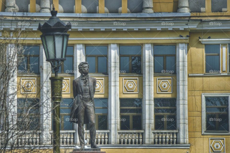 a monument to Alexander Sergeevich Pushkin against the backdrop of a beautiful architectural structure with columns and many square and rectangular figures and windows. there is a lantern nearby.