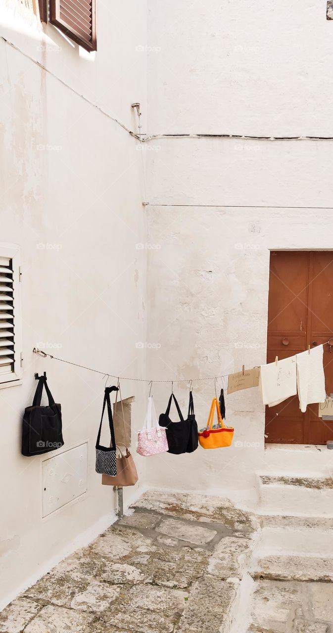 Women's handbags of different colors, sizes and styles hang on metal hooks on a clothesline in the middle of the street in the corner between houses in the city of ostuni in italy, close-up side view. Italian creative concept.