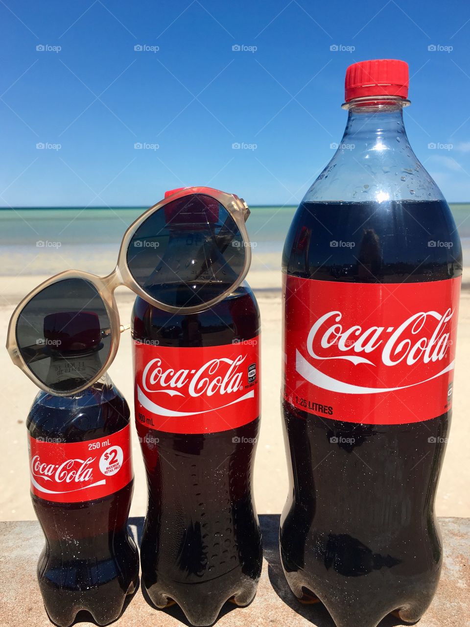 Three bottles of various sizes of Coca Cola in front of an ocean horizon suggesting a great beverage to take along on a hot day