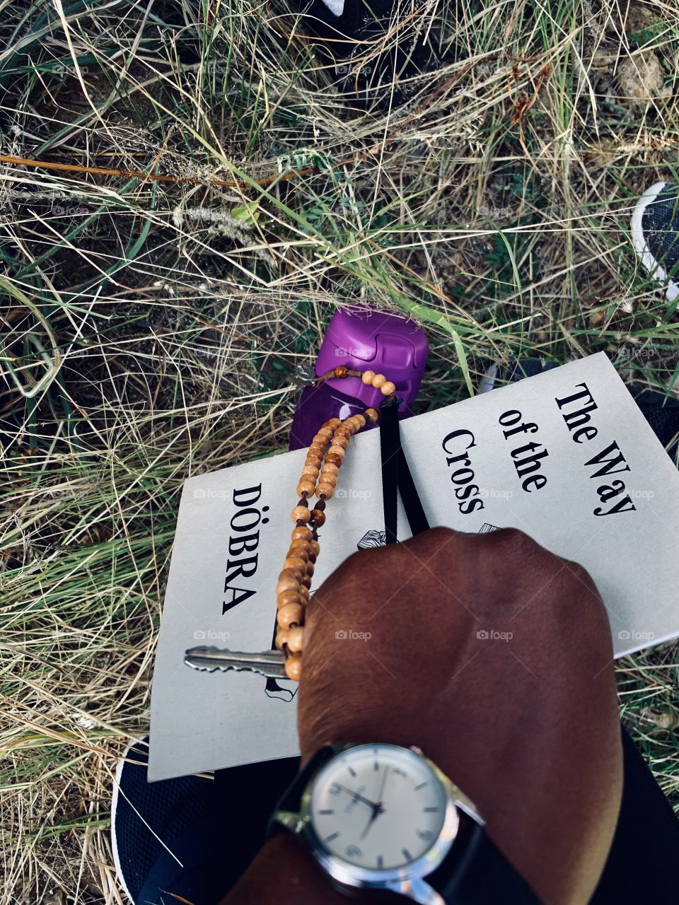 A non-Caucasian hand with a watch on the wrist holding a prayer book, a bottle of water, a rosary and keys on a pilgrimage. 