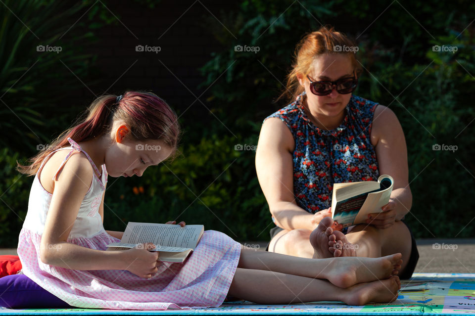 Girl and mother absorbed in reading books in the garden during sunny evening.