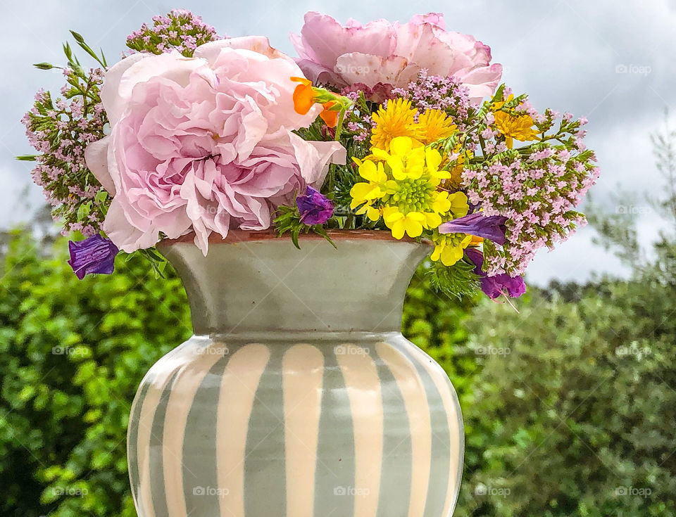 A bright and colourful selection of wild flowers sit in a cream and green ceramic pot in front of a backdrop of foliage and sky