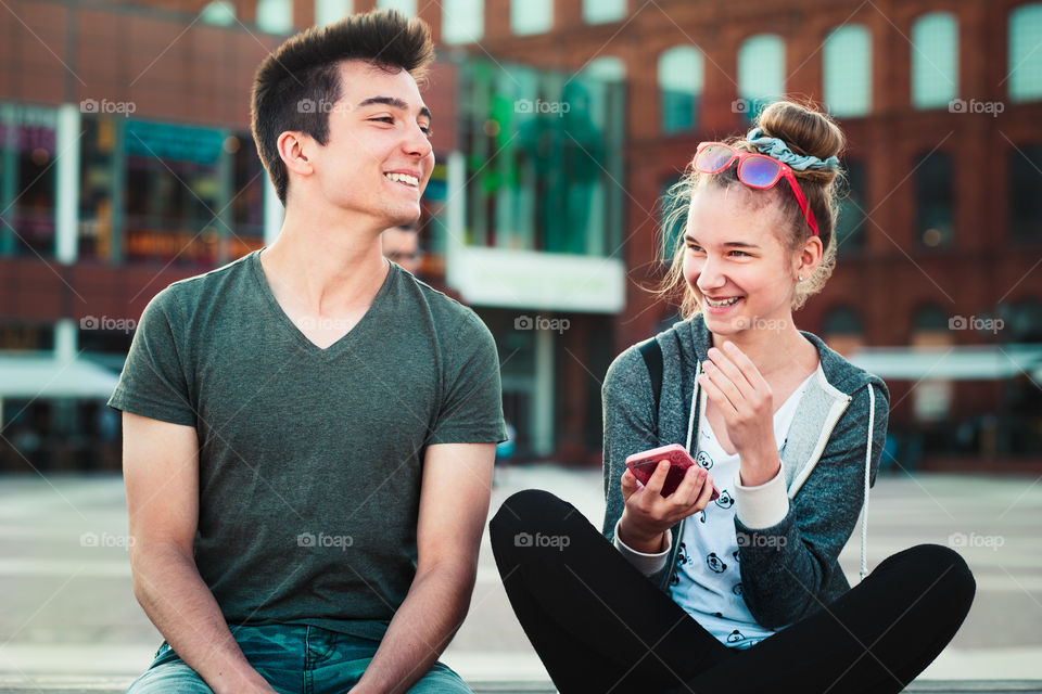 Couple of friends, teenage girl and boy,  having fun together, using smartphones,  sitting in center of town, spending time together. Real people, authentic situations